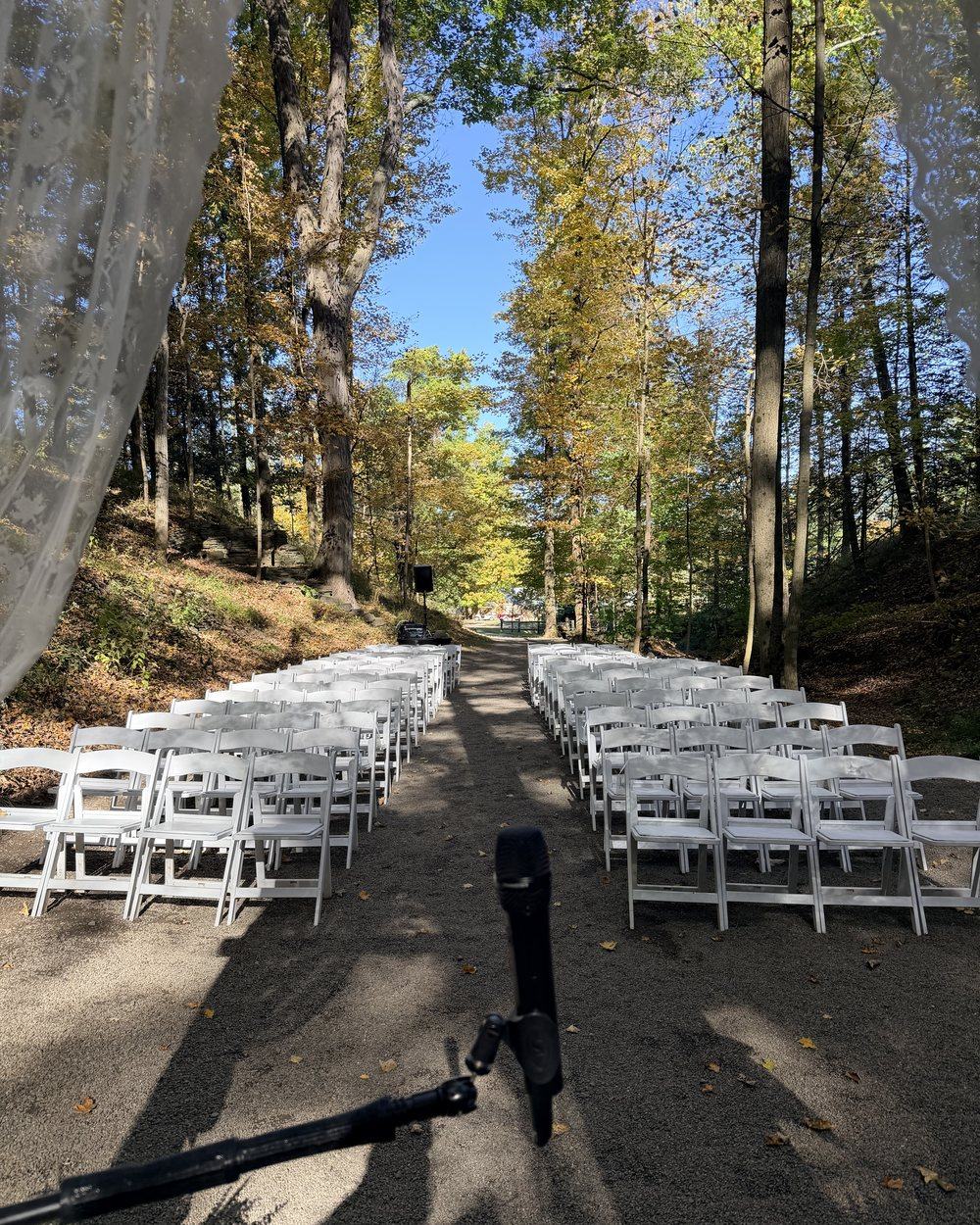 Looking back up the gravel aisle at New Park from the arch, white chairs on both sides under tall trees