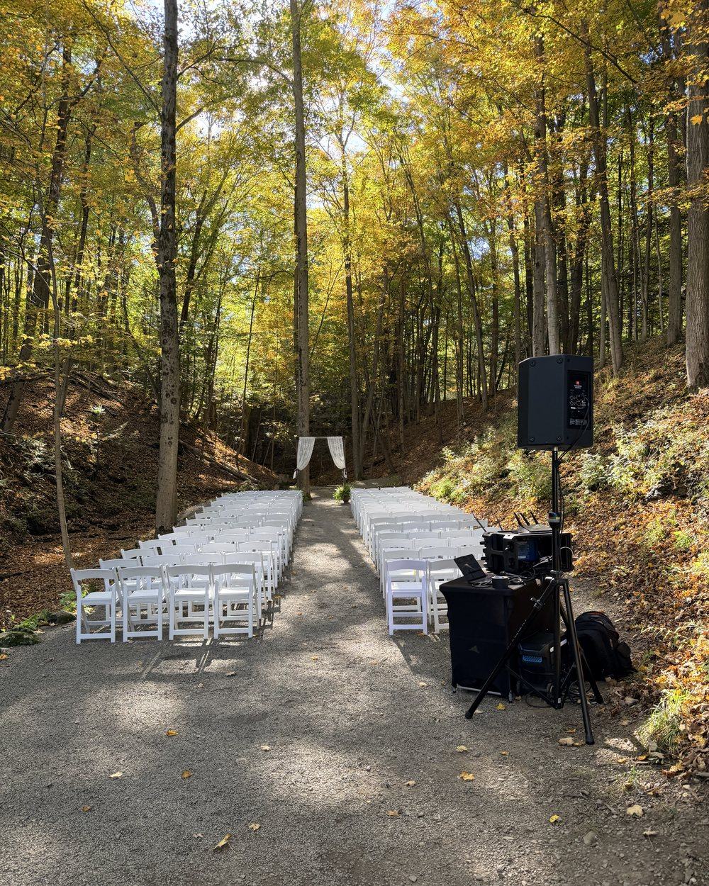 Ceremony chairs along a gravel aisle in the woods at New Park, with a draped arch at the end and a speaker on a stand to the side