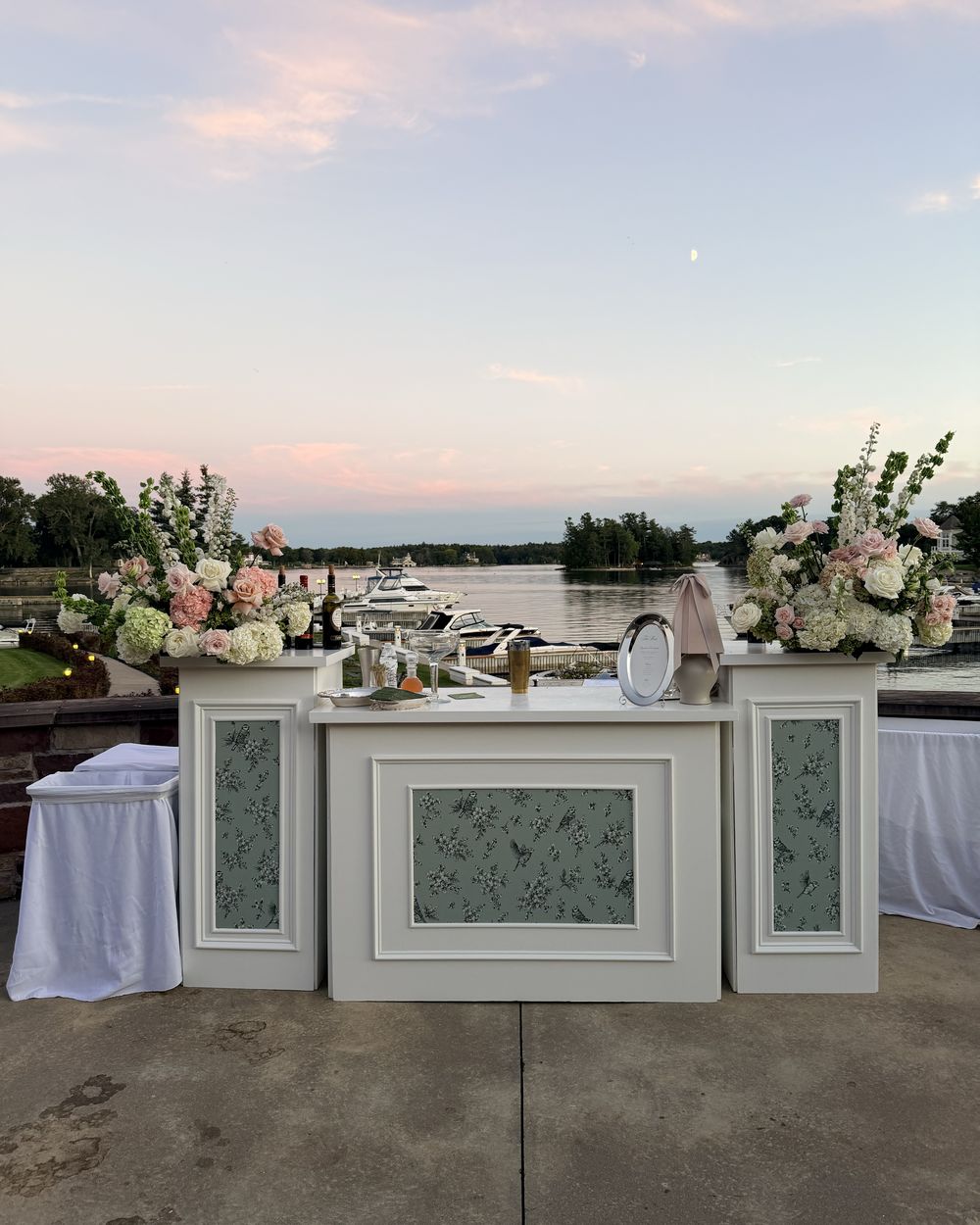 White bar with floral arrangements on the terrace above the marina and the St. Lawrence River at sunset